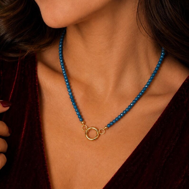 May include: A necklace featuring a strand of small, round, deep blue beads. The two ends of the beaded strand are connected to a central, circular gold-toned metal ring clasp, which serves as the focal point of the jewellery piece.