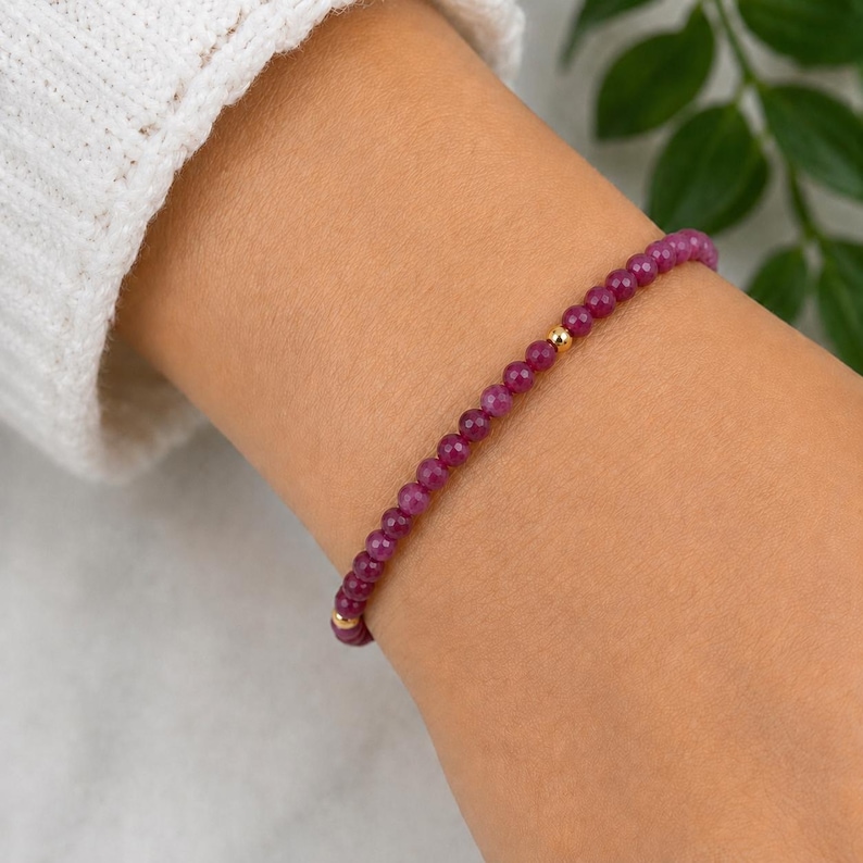 May include: A delicate beaded bracelet featuring small, round, deep magenta-colored gemstone beads strung together. The design is accented by a single, small, polished gold-toned metal bead positioned near the center of the strand.
