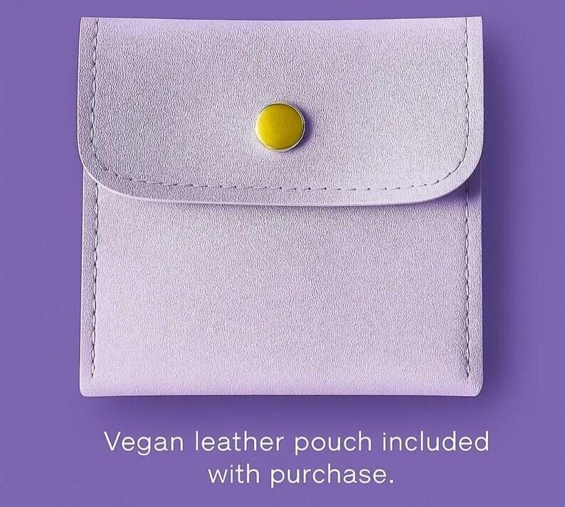 May include: A square light purple vegan leather pouch featuring a rounded flap closure secured by a single gold-tone metal snap button. The text below the pouch reads: Vegan leather pouch included with purchase.