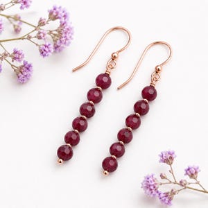 May include: A pair of dangle earrings with a rose gold finish. Each earring features a series of faceted, deep red beads strung together. The earrings are displayed on a white surface, with sprigs of purple flowers in the background.