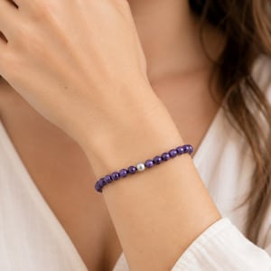 May include: A beaded bracelet featuring a series of small, round, deep purple faceted stones arranged in a continuous line, punctuated by a single, smooth, metallic silver-toned bead positioned near the center of the circular wrist accessory.
