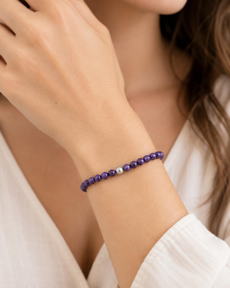 May include: A beaded bracelet featuring a series of small, round, deep purple faceted stones arranged in a continuous line, punctuated by a single, smooth, metallic silver-toned bead positioned near the center of the circular wrist accessory.