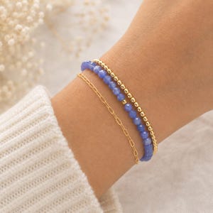 May include: A set of three bracelets consisting of one gold-tone chain link bracelet, one strand of small blue faceted gemstone beads, and one strand of small gold-tone metallic beads, all worn together on a wrist with a white ribbed sleeve.