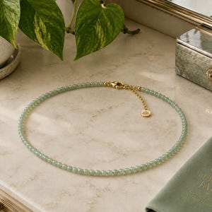 May include: A beaded necklace featuring small, round, pale green jade beads strung together. The necklace includes a gold-toned metal lobster clasp, an adjustable extender chain, and a small circular charm with a lotus flower emblem engraved on it.