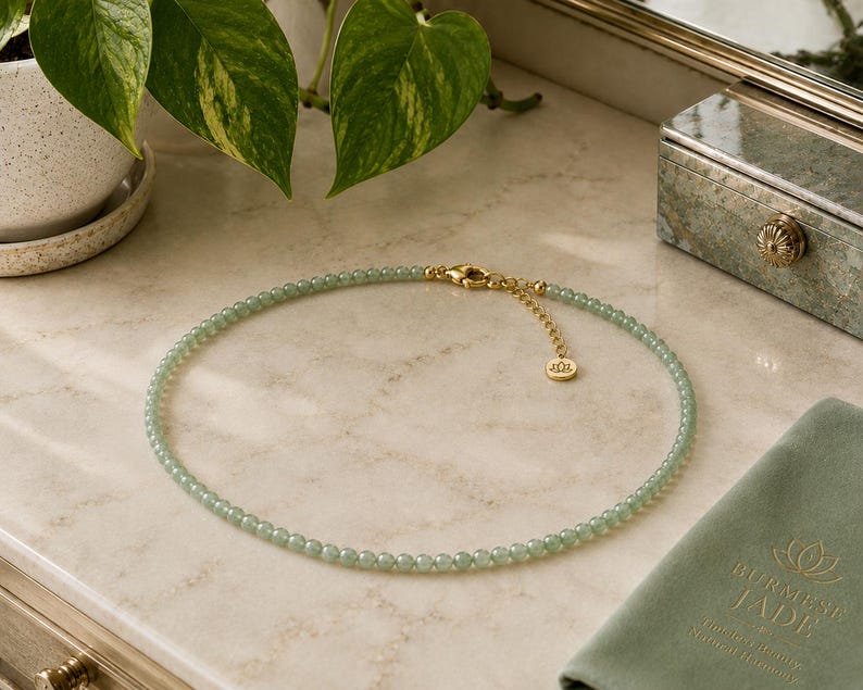 May include: A beaded necklace featuring small, round, pale green jade beads strung together. The necklace includes a gold-toned metal lobster clasp, an adjustable extender chain, and a small circular charm with a lotus flower emblem engraved on it.