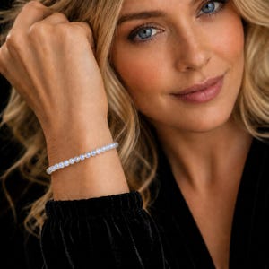 May include: A delicate bracelet composed of small, round, translucent beads with a subtle iridescent sheen. The beads are strung together in a single continuous line, creating a smooth, circular band that rests against the wrist.