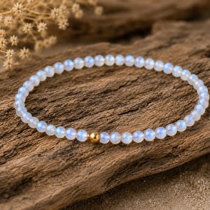 May include: A delicate bracelet composed of small, round, translucent moonstone beads with a soft blue iridescent sheen. The circular strand features a single, polished, spherical gold-toned metal bead positioned as a central accent piece.