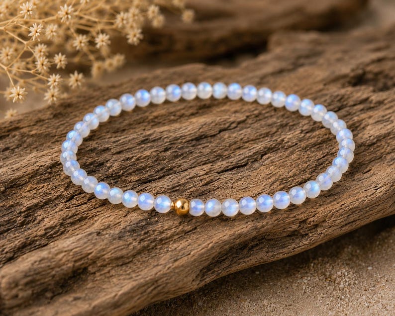 May include: A delicate bracelet composed of small, round, translucent moonstone beads with a soft blue iridescent sheen. The circular strand features a single, polished, spherical gold-toned metal bead positioned as a central accent piece.
