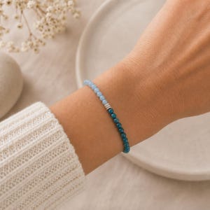 May include: A beaded bracelet featuring a gradient of small round beads transitioning from light sky blue to deep teal, accented by three small silver-toned cylindrical spacers at the center point where the two distinct color shades meet.