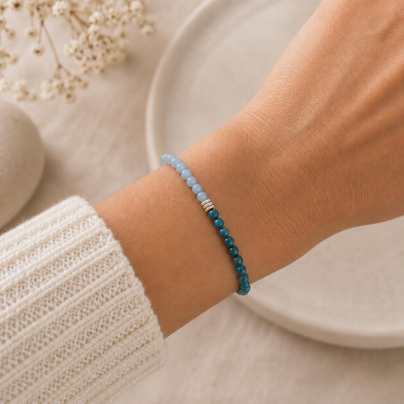May include: A beaded bracelet featuring a gradient of small round beads transitioning from light sky blue to deep teal, accented by three small silver-toned cylindrical spacers at the center point where the two distinct color shades meet.