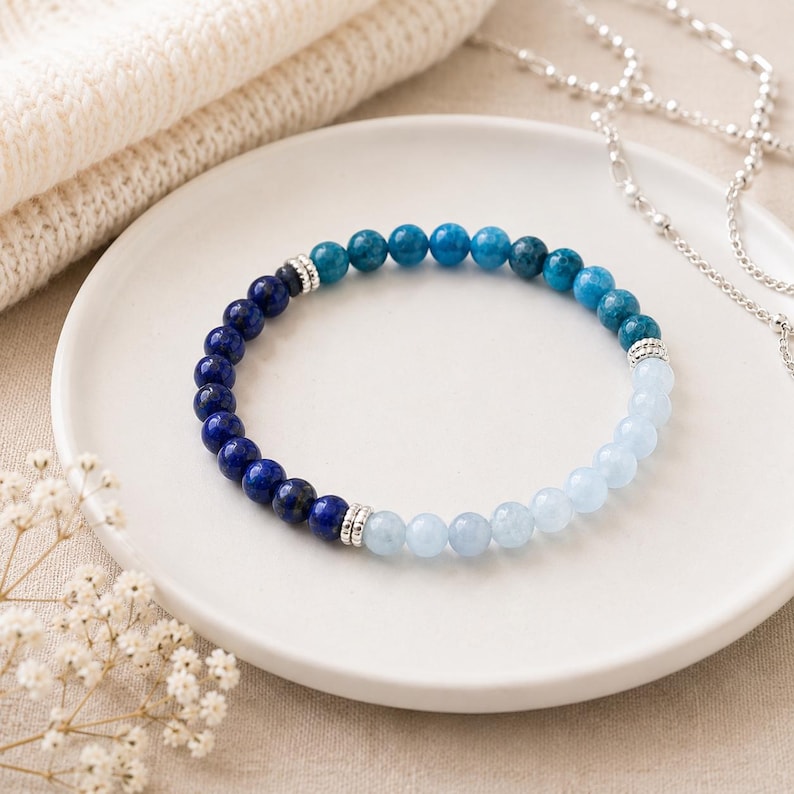 May include: A beaded bracelet featuring a gradient of round stones transitioning from deep navy blue to vibrant turquoise and light pale blue, accented with small silver-toned metal spacer beads placed at intervals along the circular strand.