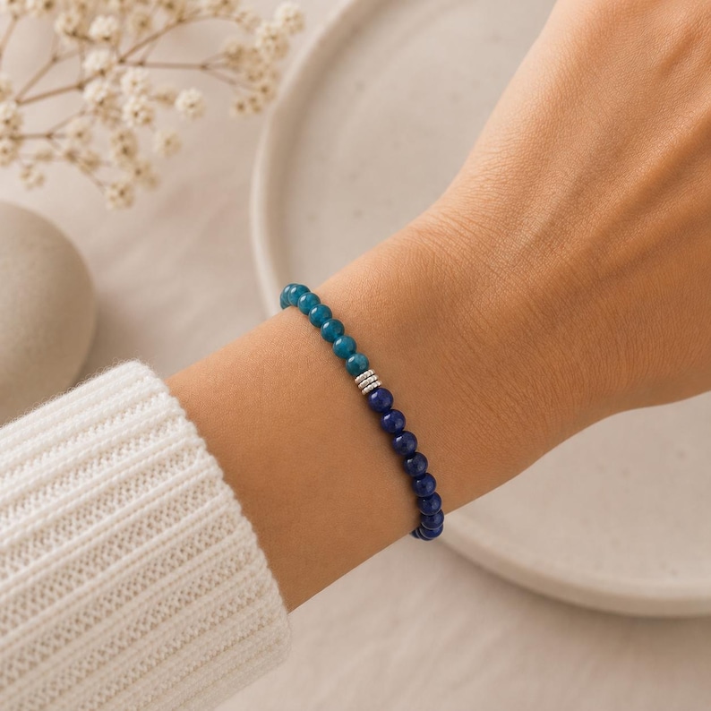 May include: A beaded bracelet featuring a gradient of small round stones transitioning from teal to deep blue, accented by three small silver-toned cylindrical spacers at the center point where the two colors meet on the elastic cord.