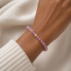 May include: A beaded bracelet featuring smooth, round, light purple gemstone beads arranged in a continuous circular pattern, accented by a single, central, textured gold-toned spherical bead that stands out against the uniform purple stones.