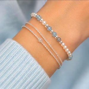 Pu&ograve; includere: Un set di tre bracciali composto da un filo di perle bianche e perline blu, una sottile catenina d'argento con un piccolo ciondolo a forma di fiore e una semplice catenina d'argento sottile, indossati al polso su un tessuto azzurro.