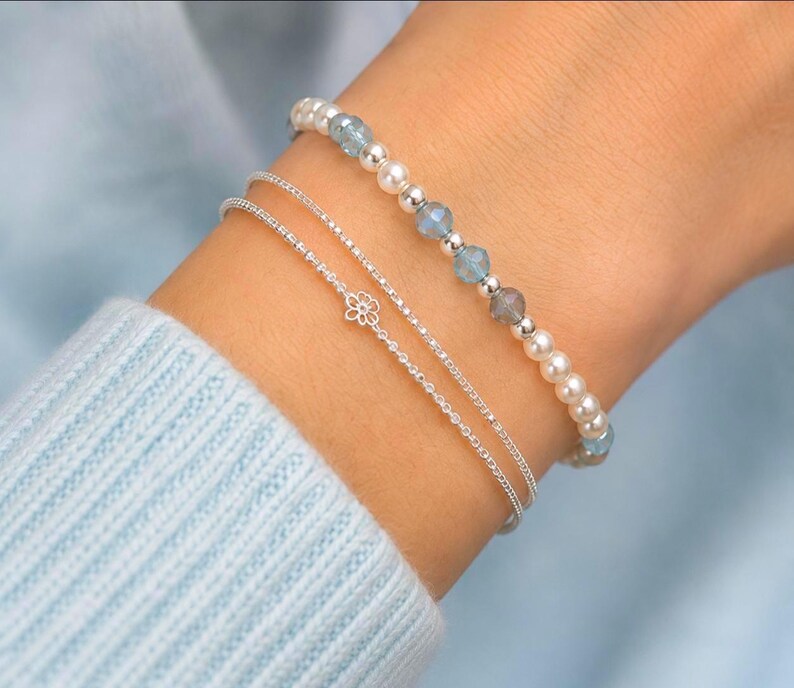 Pu&ograve; includere: Un set di tre bracciali composto da un filo di perle bianche e perline blu, una sottile catenina d'argento con un piccolo ciondolo a forma di fiore e una semplice catenina d'argento sottile, indossati al polso su un tessuto azzurro.