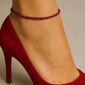 Könnte beinhalten: Ein weinrotes Perlenarmband umschließt einen Knöchel, kombiniert mit einem roten High-Heel-Schuh. Das Armband besteht aus kleinen, runden Perlen. Der Schuh hat eine spitze Zehenpartie und einen schlanken Absatz.
