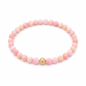 May include: A circular beaded bracelet composed of numerous smooth, round, light pink beads. A single, larger, textured gold-toned spherical bead is positioned at the bottom centre of the bracelet, contrasting with the uniform pink beads.