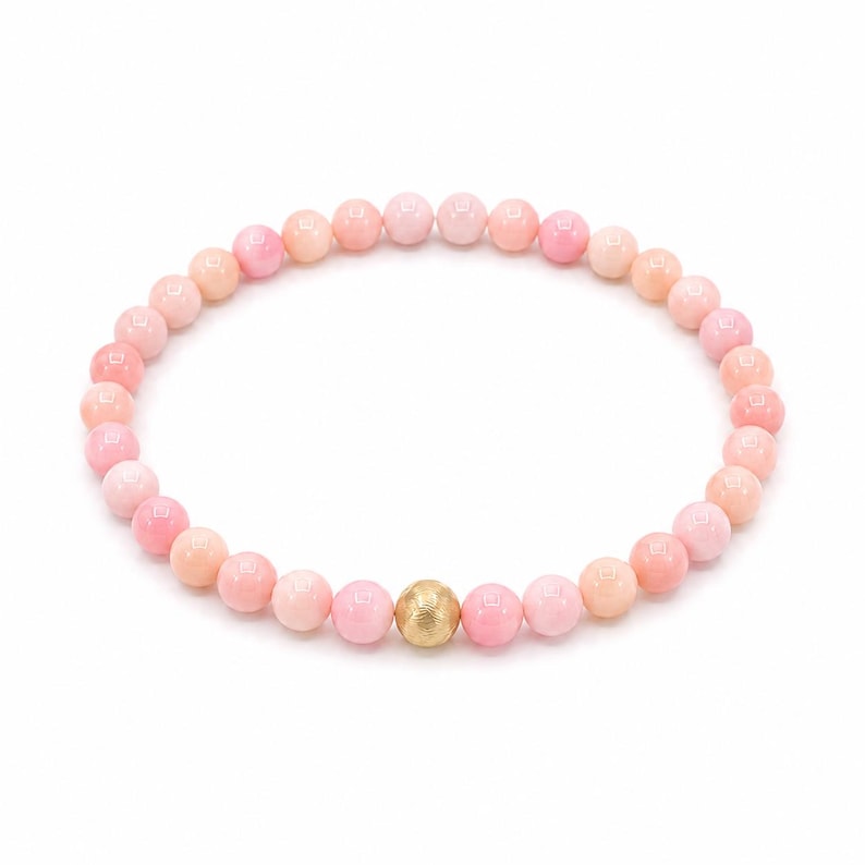 May include: A circular beaded bracelet composed of numerous smooth, round, light pink beads. A single, larger, textured gold-toned spherical bead is positioned at the bottom centre of the bracelet, contrasting with the uniform pink beads.
