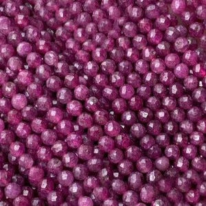May include: A large collection of small, round, faceted beads in a deep magenta-purple color. Each bead features multiple flat surfaces that catch the light, creating a textured, sparkling appearance across the entire surface of the pile.