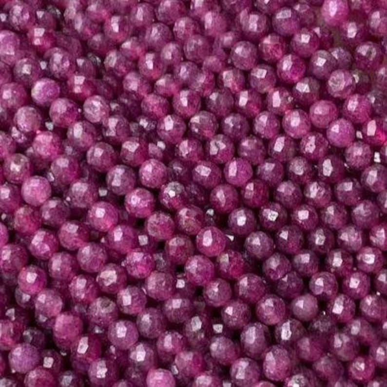 May include: A large collection of small, round, faceted beads in a deep magenta-purple color. Each bead features multiple flat surfaces that catch the light, creating a textured, sparkling appearance across the entire surface of the pile.