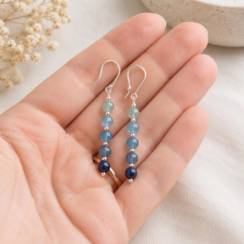 May include: A pair of dangle earrings featuring a vertical row of five round beads in a gradient of blue shades, ranging from light aquamarine to deep navy, separated by small silver-toned spacers and attached to silver-toned shepherd hook wires.