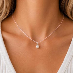 May include: A delicate necklace featuring a single, round, white pearl pendant suspended from a thin, light pink cord. The pearl is attached to the cord with a small, simple silver-toned metal bail, creating a minimalist and clean appearance.