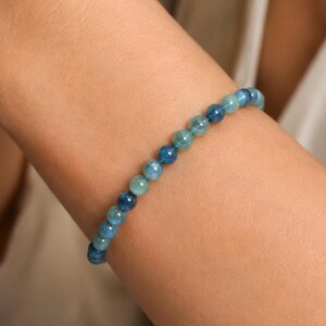 May include: A beaded bracelet featuring small, round, polished stones in varying shades of blue, ranging from light aquamarine to deep navy. The beads are strung together in a continuous line, creating a smooth, uniform appearance around the wrist.