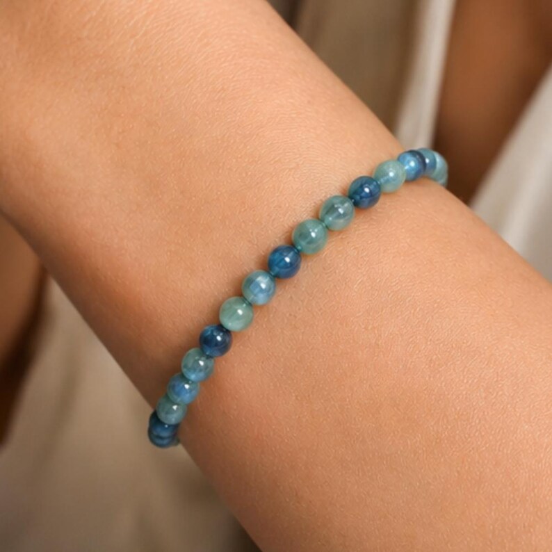 May include: A beaded bracelet featuring small, round, polished stones in varying shades of blue, ranging from light aquamarine to deep navy. The beads are strung together in a continuous line, creating a smooth, uniform appearance around the wrist.