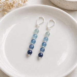 May include: A pair of drop earrings featuring a vertical arrangement of five faceted beads in an ombre color gradient, transitioning from light blue at the top to dark navy blue at the bottom, separated by small silver-toned metal spacers.