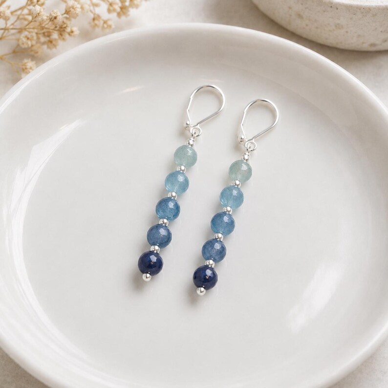 May include: A pair of drop earrings featuring a vertical arrangement of five faceted beads in an ombre color gradient, transitioning from light blue at the top to dark navy blue at the bottom, separated by small silver-toned metal spacers.