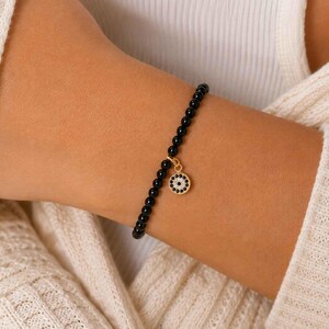 May include: A beaded bracelet featuring small, smooth, round black beads strung together. A circular gold-toned charm hangs from the center, displaying a detailed eye motif crafted with tiny black and white stones set in a gold-colored metal frame.