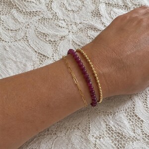 May include: A set of three bracelets worn together. One is a thin gold paperclip chain, the second is a strand of small, faceted deep magenta gemstone beads, and the third is a delicate gold beaded chain with small, round, polished metal spheres.