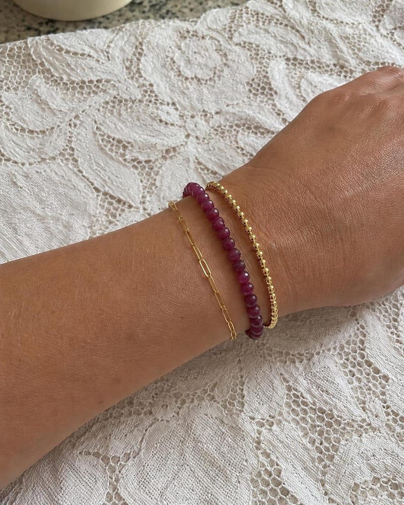 May include: A set of three bracelets worn together. One is a thin gold paperclip chain, the second is a strand of small, faceted deep magenta gemstone beads, and the third is a delicate gold beaded chain with small, round, polished metal spheres.