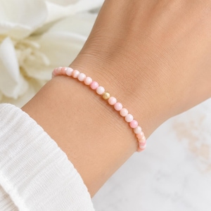 May include: A delicate bracelet composed of small, round, light pink beads arranged in a continuous strand. A single, smooth, metallic gold-toned bead is positioned as a central accent among the pink beads, creating a subtle contrast in colour and texture.