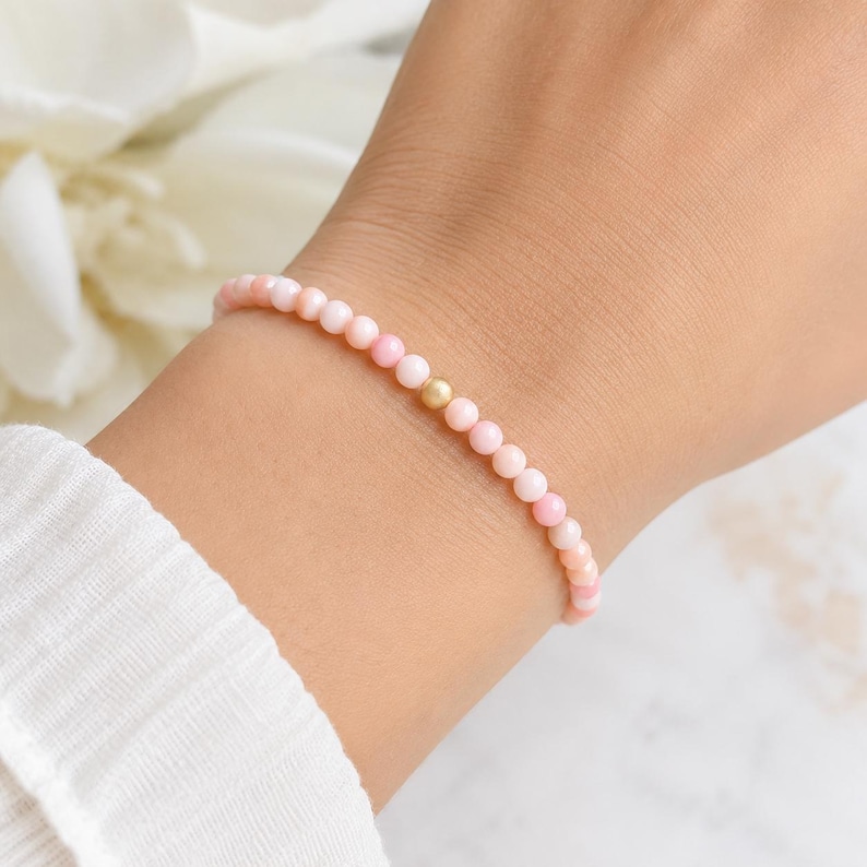 May include: A delicate bracelet composed of small, round, light pink beads arranged in a continuous strand. A single, smooth, metallic gold-toned bead is positioned as a central accent among the pink beads, creating a subtle contrast in colour and texture.