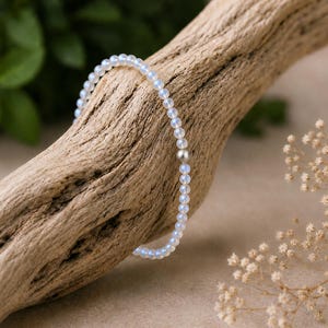 May include: A delicate beaded bracelet featuring small, translucent, iridescent round beads with a soft blue glow. The bracelet includes a single, smooth, matte-finish silver-toned metal bead positioned as a central accent among the light-reflecting stones.
