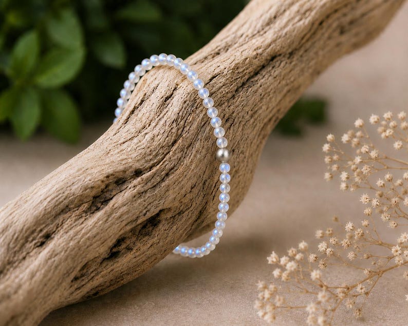 May include: A delicate beaded bracelet featuring small, translucent, iridescent round beads with a soft blue glow. The bracelet includes a single, smooth, matte-finish silver-toned metal bead positioned as a central accent among the light-reflecting stones.