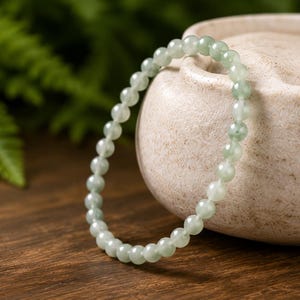 May include: A circular bracelet composed of small, smooth, translucent pale green beads. The beads exhibit subtle variations in color, ranging from light mint to soft sage, and are strung together in a continuous loop to form a simple, uniform band.