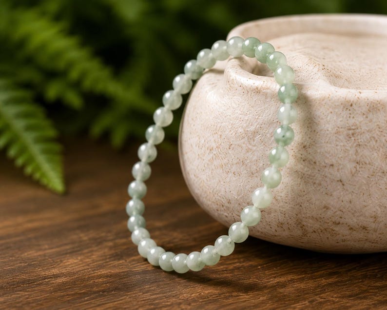 May include: A circular bracelet composed of small, smooth, translucent pale green beads. The beads exhibit subtle variations in color, ranging from light mint to soft sage, and are strung together in a continuous loop to form a simple, uniform band.