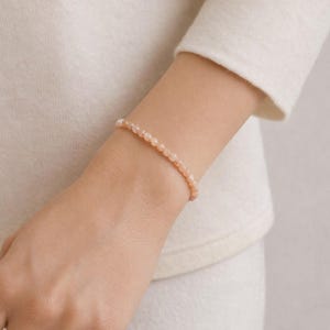 May include: A bracelet made of small, peach-coloured beads. The bracelet is worn on a wrist, set against a cream-coloured long-sleeved garment. The beads are evenly sized and strung together. The bracelet is likely to be 16-18 cm in length.