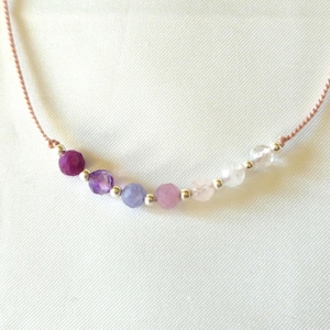 May include: A delicate necklace featuring a thin tan cord strung with a gradient of seven faceted gemstone beads in shades of deep purple, lavender, light blue, soft pink, and clear white, separated by small, round, polished silver-tone metal beads.