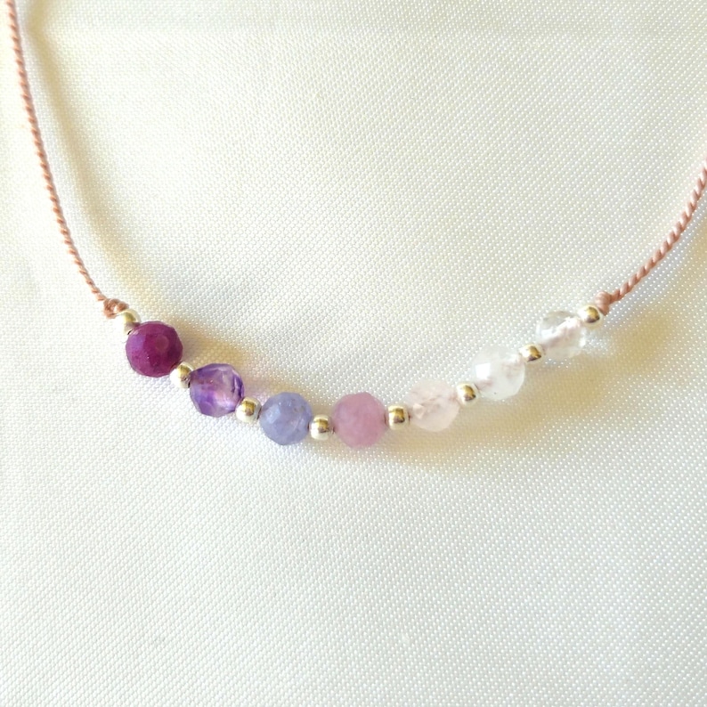 May include: A delicate necklace featuring a thin tan cord strung with a gradient of seven faceted gemstone beads in shades of deep purple, lavender, light blue, soft pink, and clear white, separated by small, round, polished silver-tone metal beads.