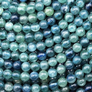 May include: A collection of round, polished kyanite beads featuring a spectrum of blue and green hues. Each bead displays natural internal inclusions and a glossy, reflective surface, creating a dense, textured arrangement of gemstone spheres.