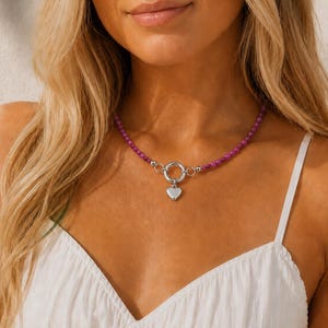May include: A necklace featuring a strand of small, round, vibrant purple beads. The necklace is secured with a silver-toned circular toggle clasp at the centre, from which a smooth, polished silver-toned heart-shaped pendant hangs.