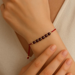 Peut inclure: Bracelet en fil rouge orné de sept perles rouge foncé et de petits détails dorés. Le bracelet est réglable grâce à un nœud coulissant. Un bijou simple et élégant.