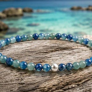 May include: A beaded bracelet featuring a continuous strand of small, polished, round blue and teal gemstone beads. The bracelet is accented with a single, central, textured silver-toned metal bead that stands out among the smooth, translucent stones.