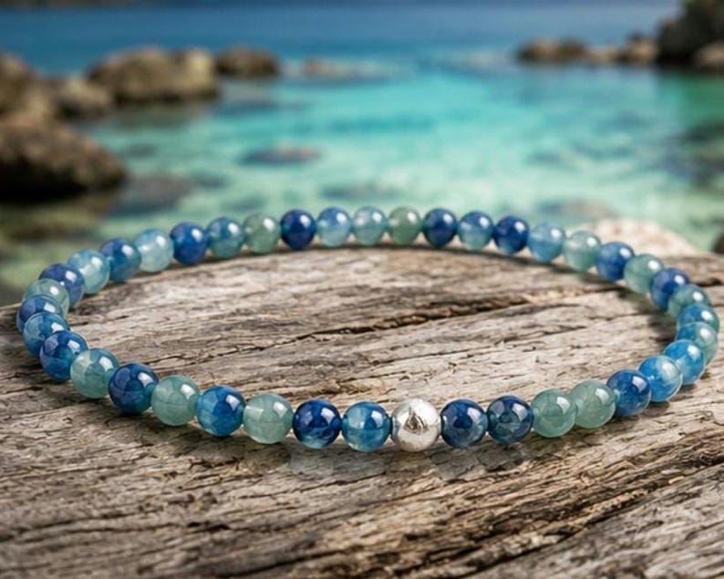 May include: A beaded bracelet featuring a continuous strand of small, polished, round blue and teal gemstone beads. The bracelet is accented with a single, central, textured silver-toned metal bead that stands out among the smooth, translucent stones.
