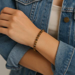 Peut inclure: Un bracelet composé de petites perles rondes en pierre œil-de-tigre marron, alternées avec de minuscules entretoises métalliques dorées. Le bracelet est porté au poignet, mettant en valeur les tons marron terreux des perles en pierre naturelle.