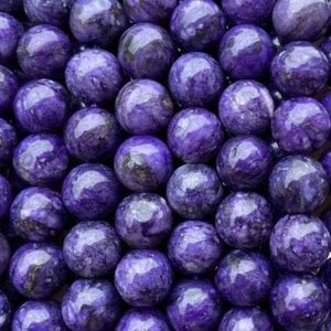 May include: A collection of polished, spherical beads made of purple charoite stone. Each bead features a smooth, glossy surface with swirling patterns of deep violet, lavender, and dark black inclusions, creating a marbled appearance throughout.