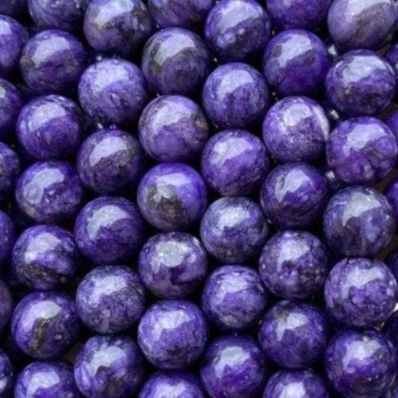 May include: A collection of polished, spherical beads made of purple charoite stone. Each bead features a smooth, glossy surface with swirling patterns of deep violet, lavender, and dark black inclusions, creating a marbled appearance throughout.
